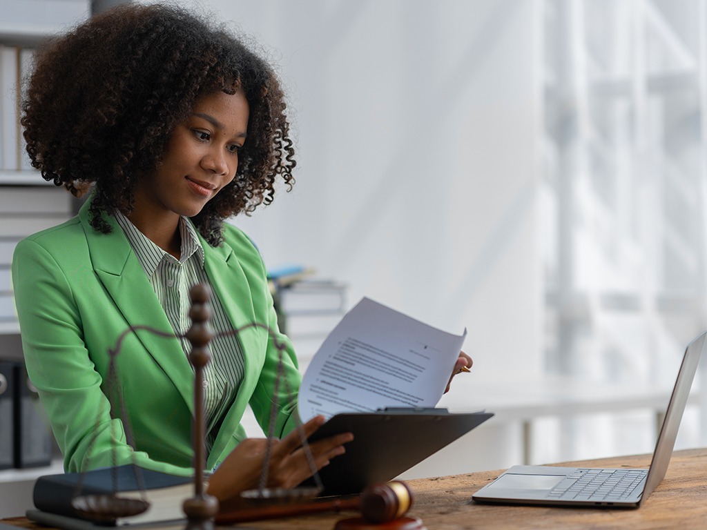A woman in a green blazer reviews documents at a desk, appearing focused and professional. A laptop, gavel, and balanced scale are on the desk.