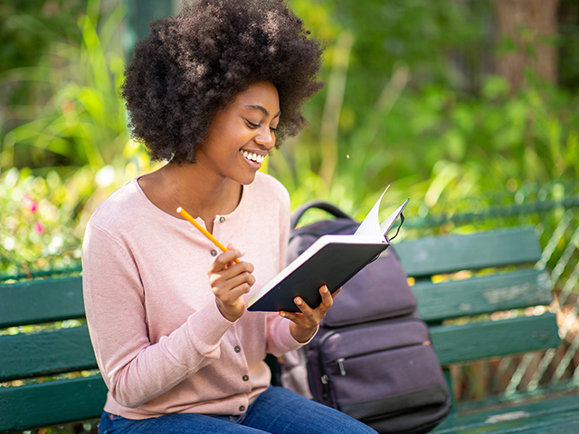 young smiling african american woman sitting outside writing in a notebook