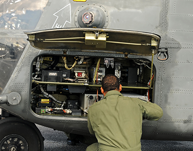 A military engineer in an army green jumpsuit repairs a helicopter engine, focused and facing away from the camera.