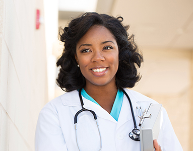 A smiling black female medical student, dressed in a white coat, stands outside a building