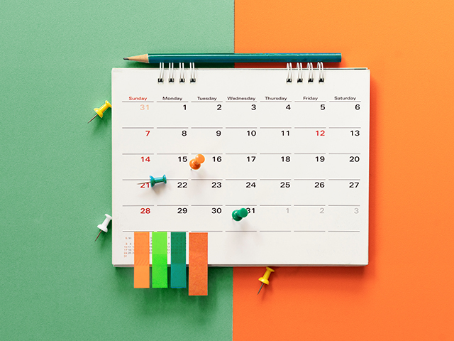 A white calendar set against an orange and green backdrop, adorned with sticky tabs and push pins for notes.