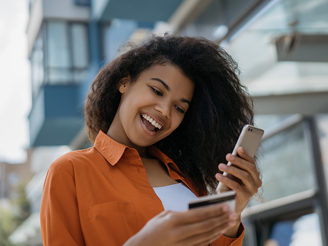 smiling young woman in orange blouse holding her phone in one hand and a credit card in the other