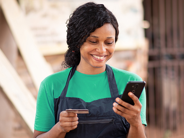 smiling woman in black apron holding her phone in one hand and a credit card in the other