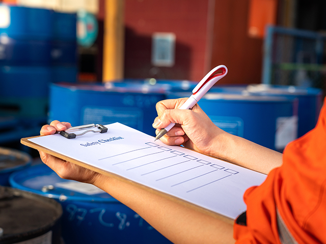 Safety inspection worker holding a pen and clipboard with a checklist document attached, a blurred background of chemical storage area