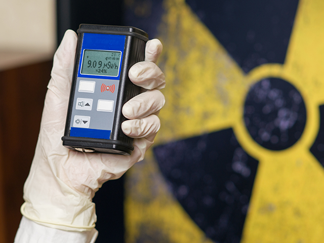 Person wearing white gloves holding a Geiger counter with radioactive materials in the background