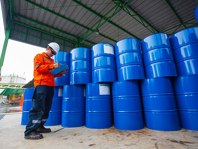 A male inspection worker holding a pen and clipboard, looking at a stock of oil drum barrels