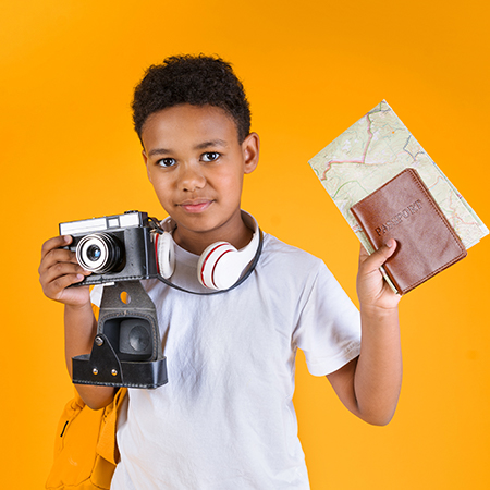Smiling traveling African American little boy with backpack, holding vintage camera, passport, and map, infront of orange background
