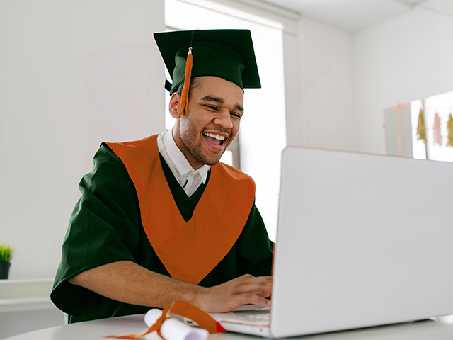 Happy male student in graduation dress looking at laptop 