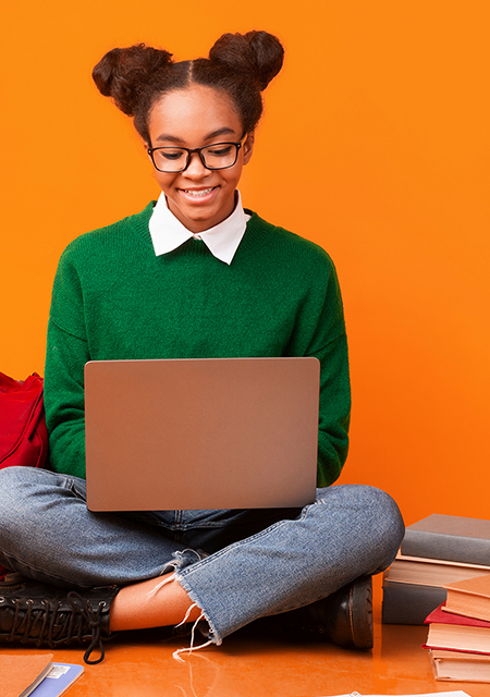 Smiling African American female student in a green shirt seated cross-legged on the floor with a laptop, next to a red backpack and a small stack of books, against an orange background