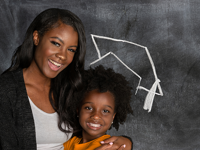 African American mother and child daughter standing in front of a black chalkboard. A chalk drawing of a graduation cap is placed over the daughter's head.