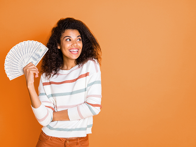 smiling young woman holding big fan of money in hands in front of orange color background