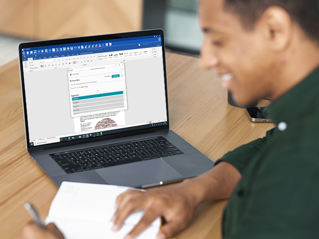 Young African American male college student takes notes in notebook, sitting at desk with laptop open