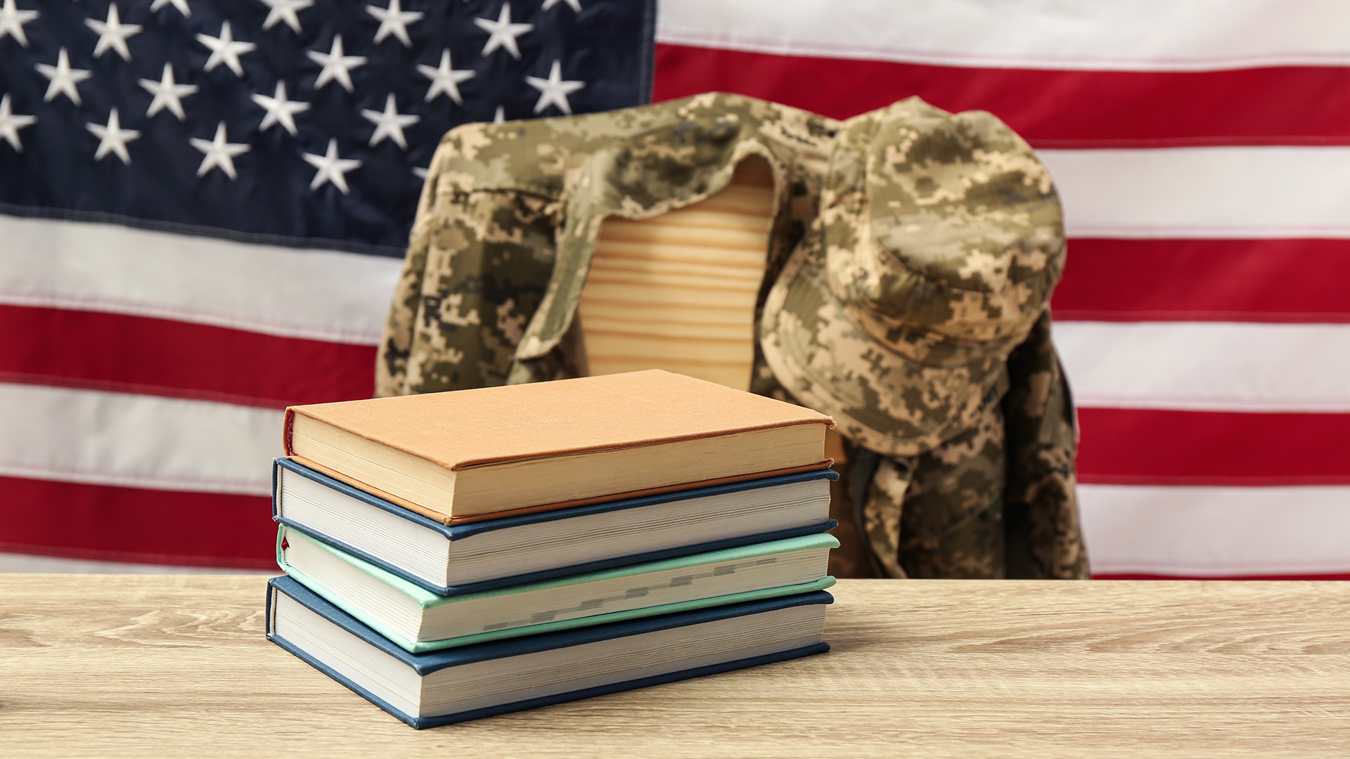 A desk with a stack of books sitting on top. Behind the desk is a chair with a military jacket draped over its back. In the background is the American flag.