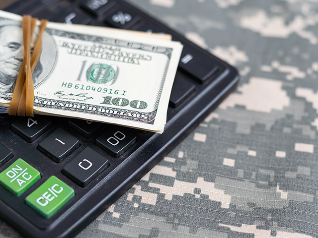 Stack of money secured with a rubber band resting on a calculator, both placed atop a camouflage cloth.